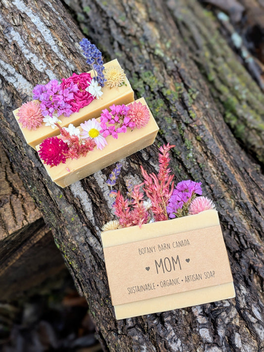 Three soap bars with floral toppings on a tree trunk, featuring a label for 'Mom' from Botany Barn Canada.