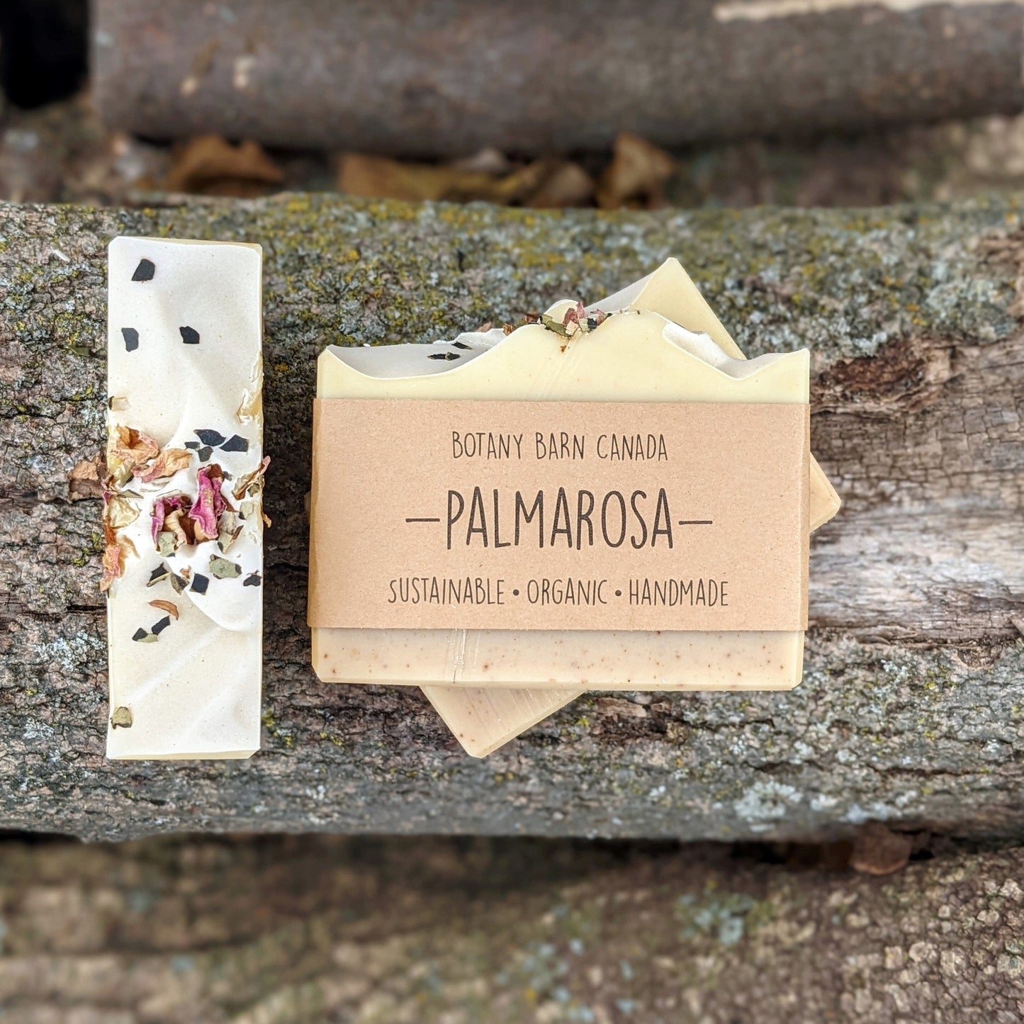 Bars of handmade artisan soap, top and front view. The soap is pale pink and yellow in colour with green and pink petals on top. A brown label reads the name of the company, Botany Barn Canada and the name of the soap, Palmarosa.