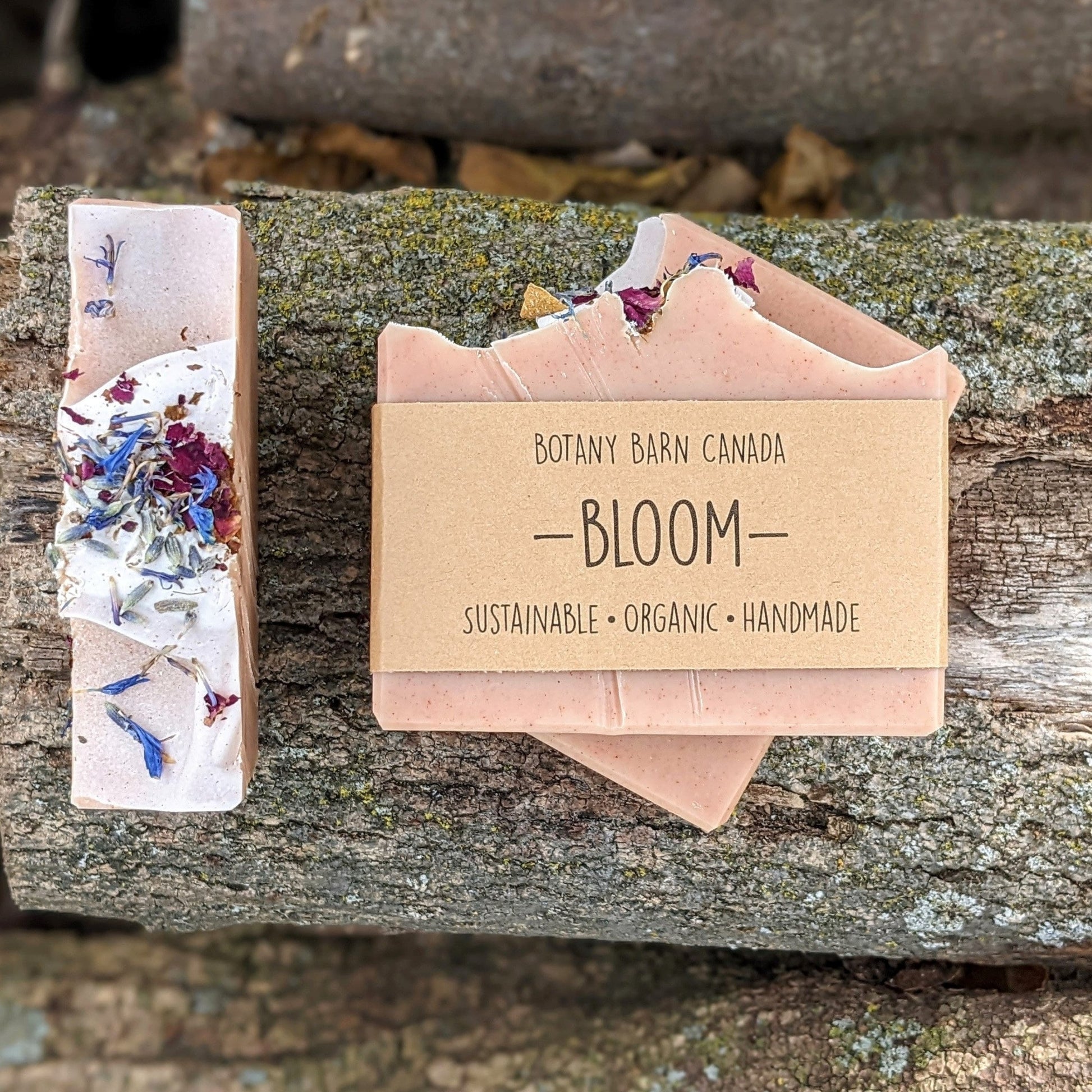 Bars of handmade artisan soap, top and front view. The soap is dusty pink in colour and decorated with brightly coloured flower petals on top. A brown label reads the name of the company, Botany Barn Canada and the name of the soap, Bloom.