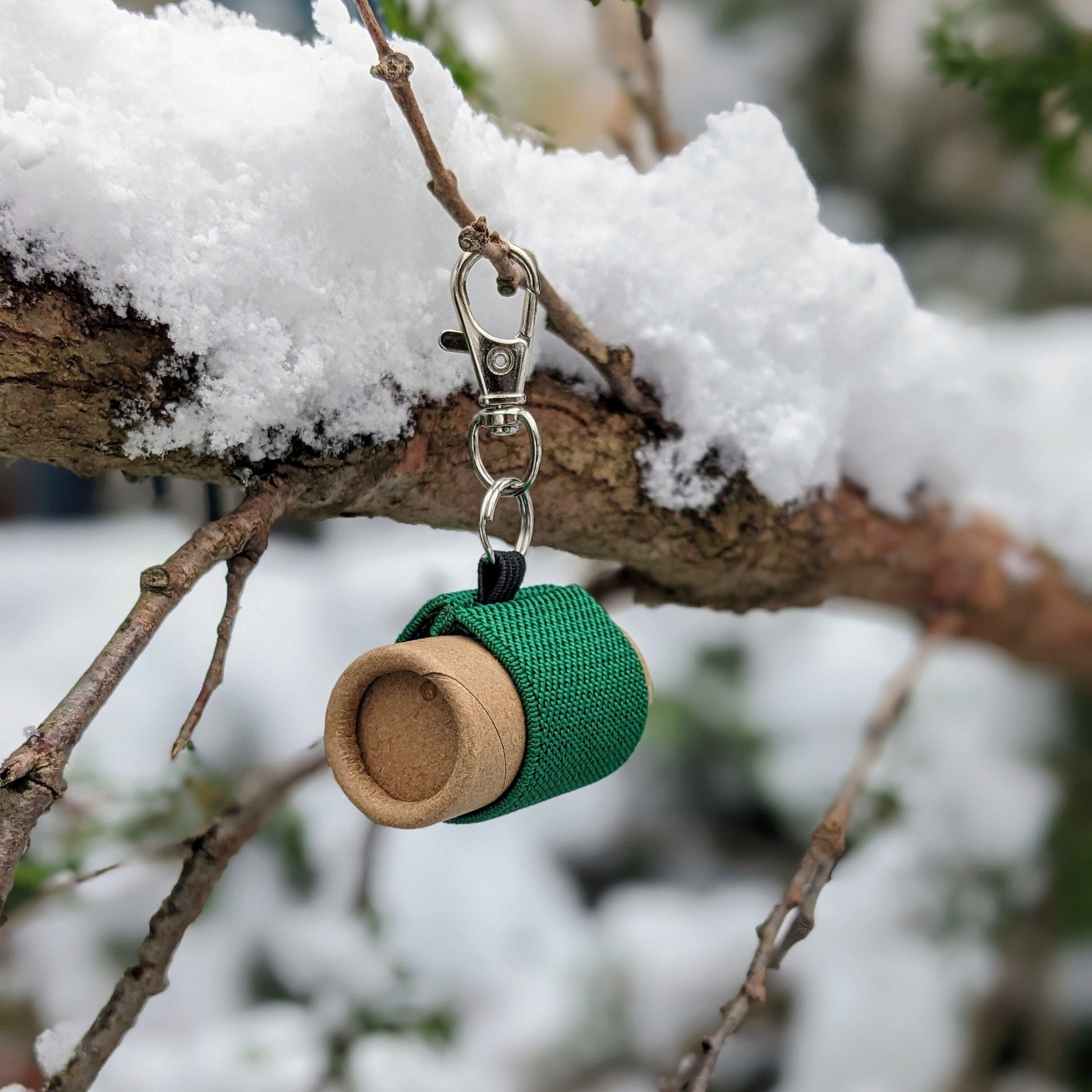 Green lip balm holder with lip balm, attached by keychain to a snow-covered branch