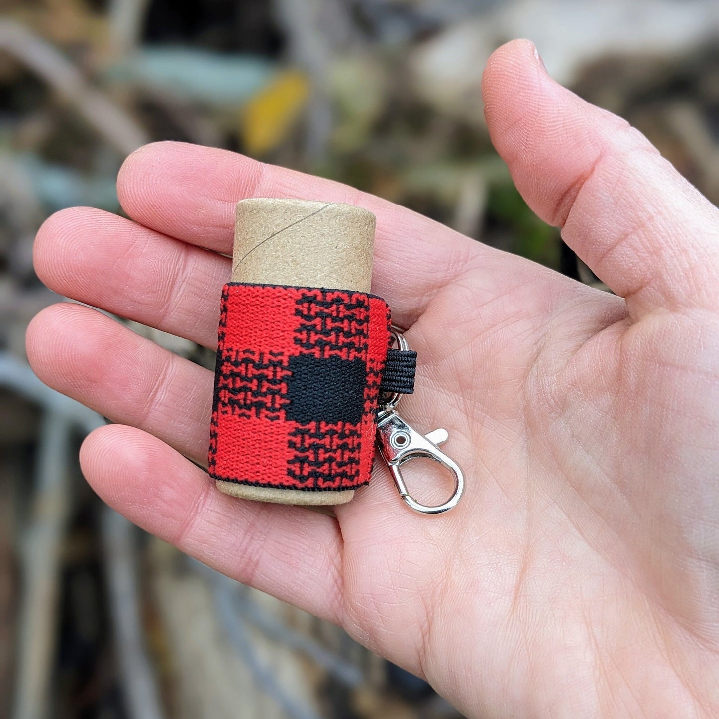Red and black checkered elastic lip balm holder with keychain, around a lip balm tube. Held in a hand with a blurred natural background