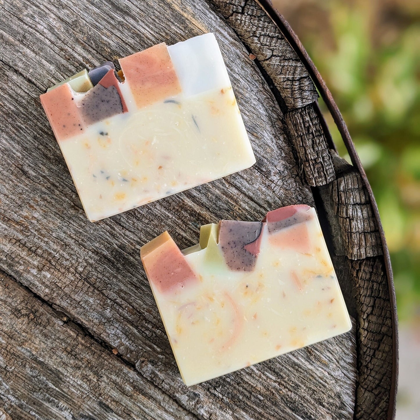 Two colourful soap bars on a wooden surface with a blurred natural background